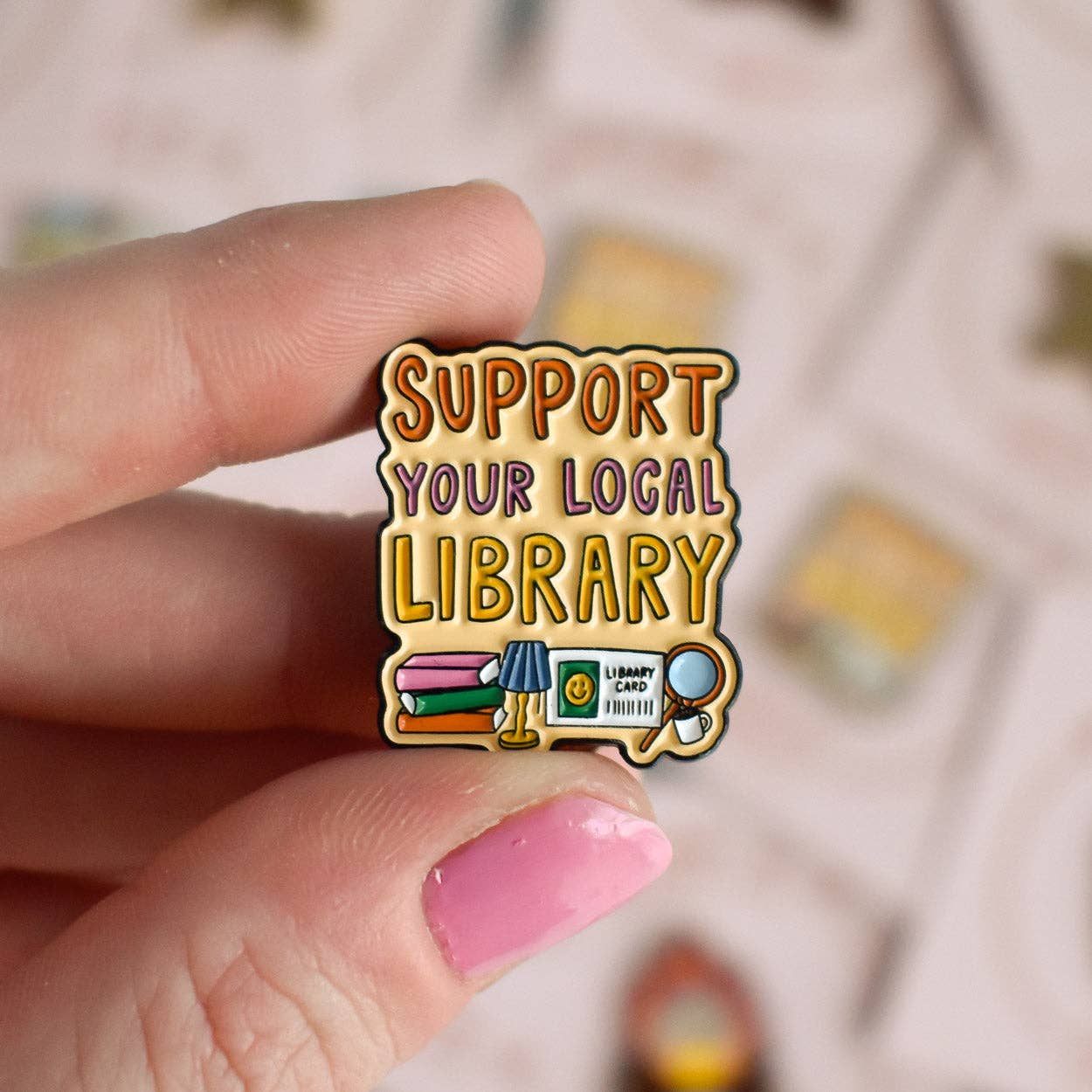 Pin with 'Support Your Local Library' text held by a hand with pink nail polish.