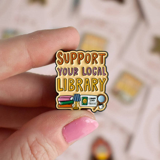 Pin with 'Support Your Local Library' text held by a hand with pink nail polish.