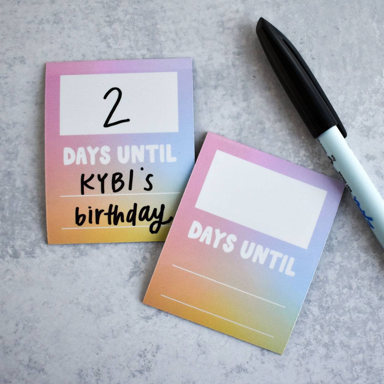 Gradient notepads with '2 days until KYBI's birthday' text and a pen on a gray surface